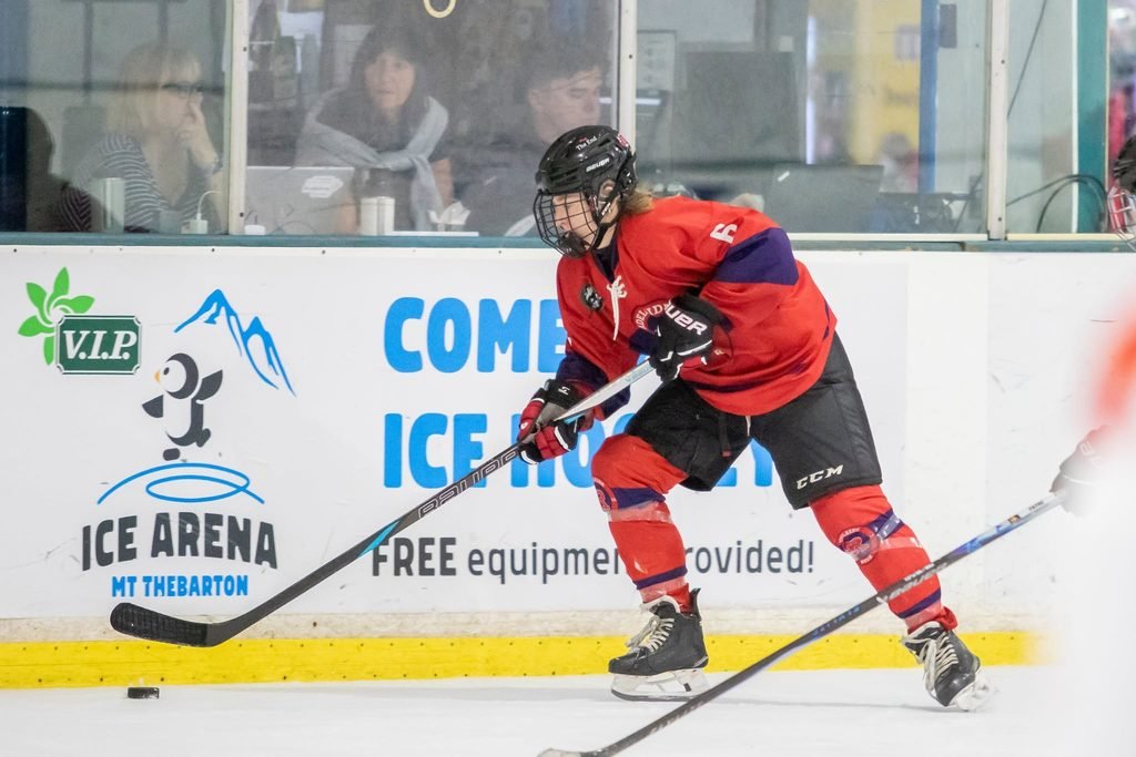 Adelaide Rush defender Tiffany Venning wearing a red, purple, and black hockey jersey with the number 6, controlling the puck along the ice rink boards next to an Ice Arena sign.