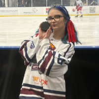 Headshot of Alma Korajac, TikTok Manager and reporter for 6ix Dub Hockey, wearing glasses and a white ice hockey jersey while holding up a puck next to her cheek at an ice rink.
