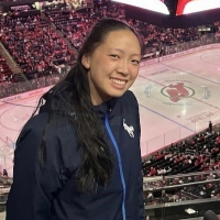 Headshot of Isabel Lay, Melbourne Ice reporter for 6ix Dub Hockey, smiling in a navy blue jacket with an ice hockey rink visible in the background stadium.
